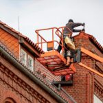 Roofer in a cherry picker conducting maintenance on a red brick building with a tiled roof.